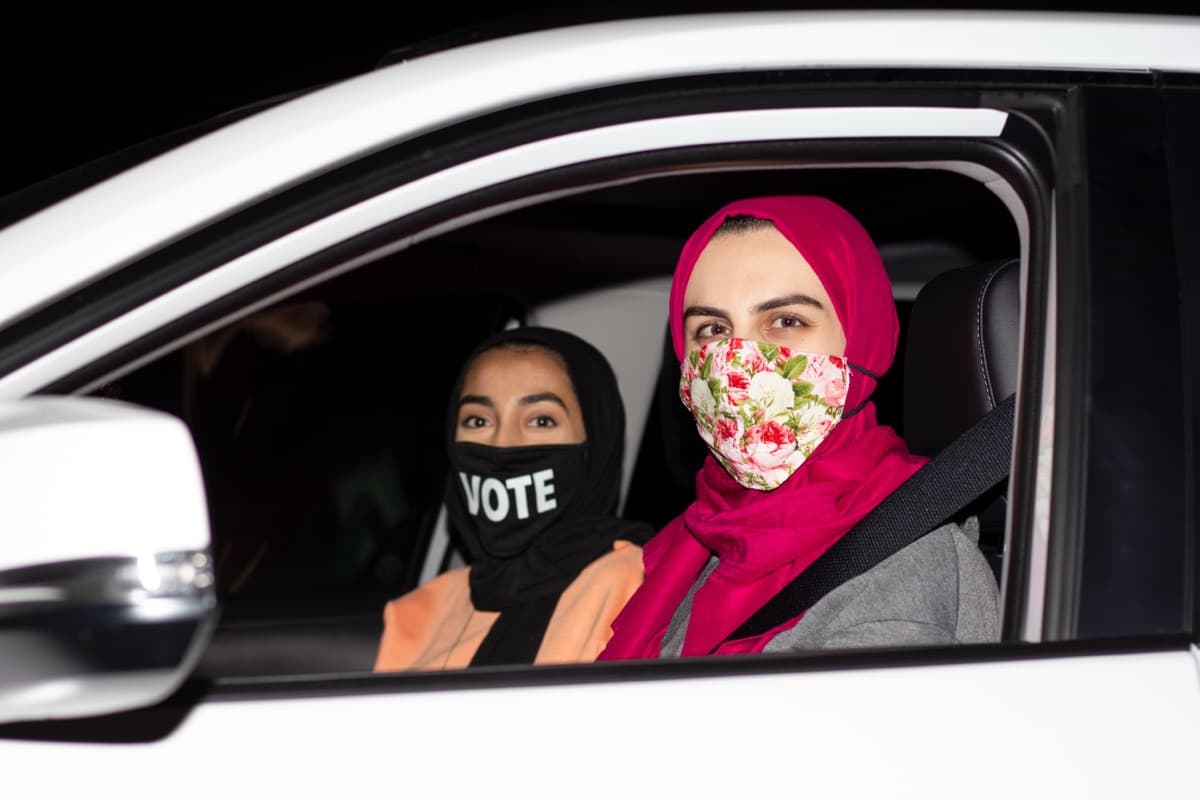 Two women in a car, one wearing a VOTE mask, at a drive-in get-out-the-vote event.