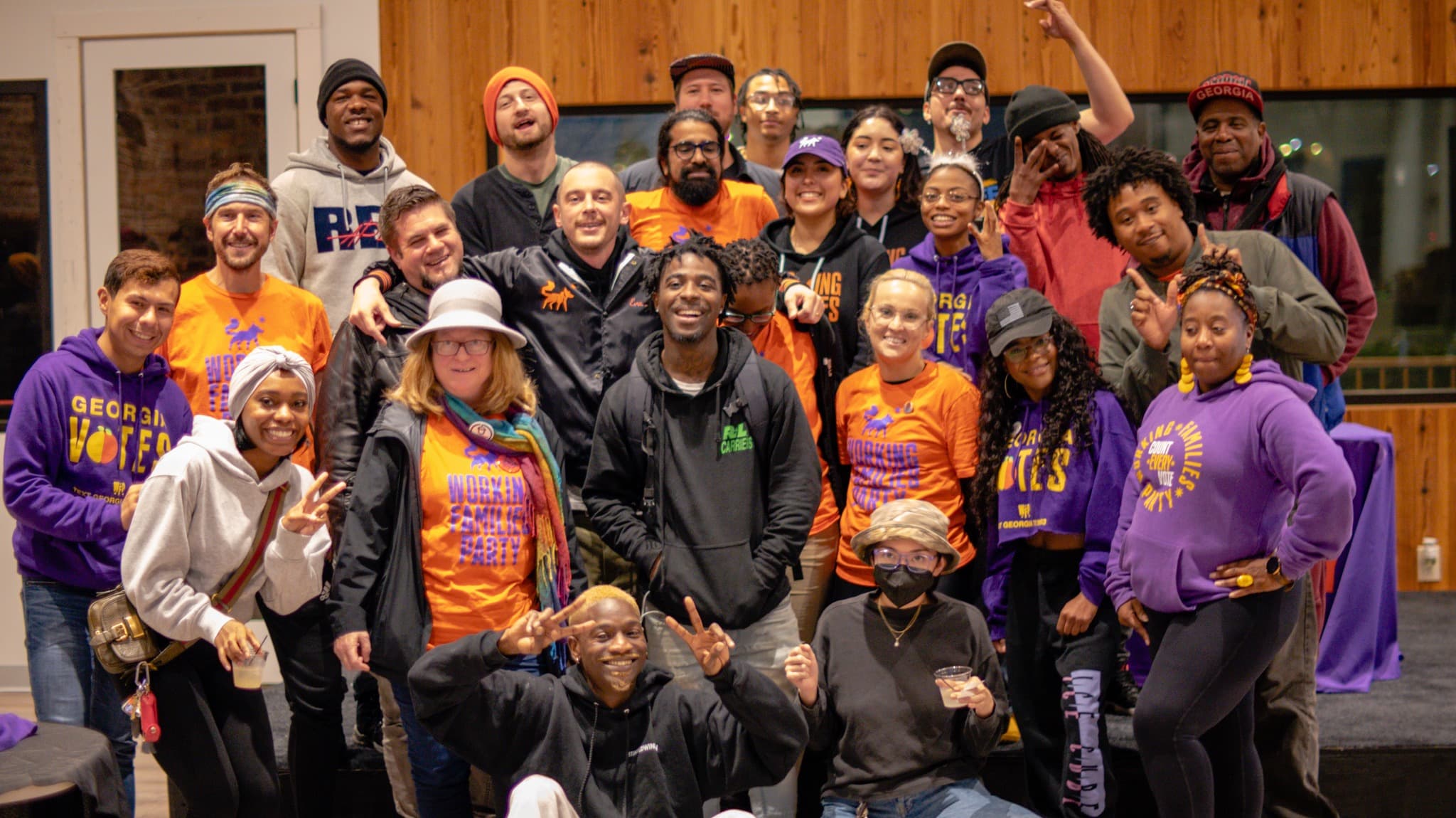 About 30 Georgia Working Families Party members and volunteers in orange WFP shirts and purple ‘Georgia Votes’ hoodies pose together indoors after the December 2022 runoff
