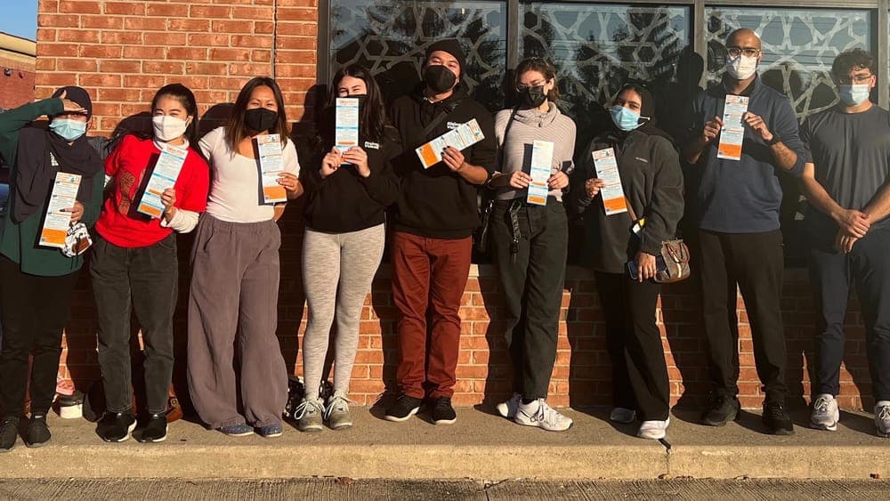 Rising Voices volunteer canvassers in masks hold up campaign literature in front of a brick wall in Macomb County.
