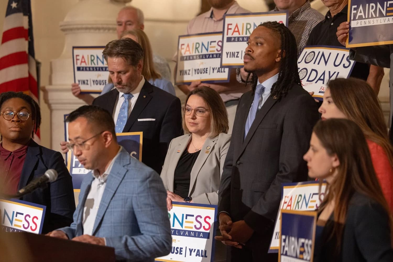 Supporters hold Fairness Pennsylvania signs behind speakers at a news conference in the Pennsylvania Capitol.