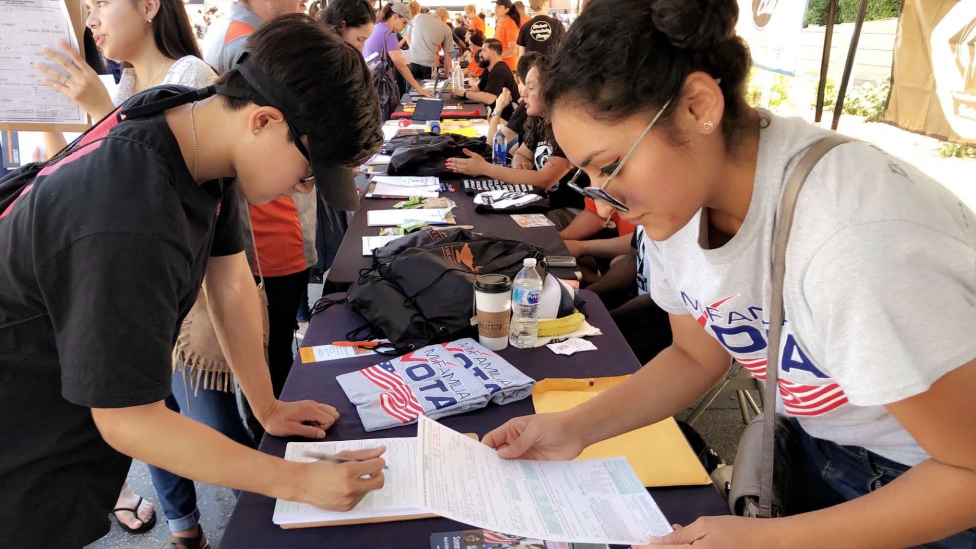 Mi Familia Vota volunteers help people fill out voter registration forms at a table.