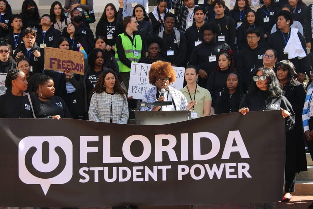 A speaker addresses a crowd of young people holding signs behind a Florida Student Power banner.