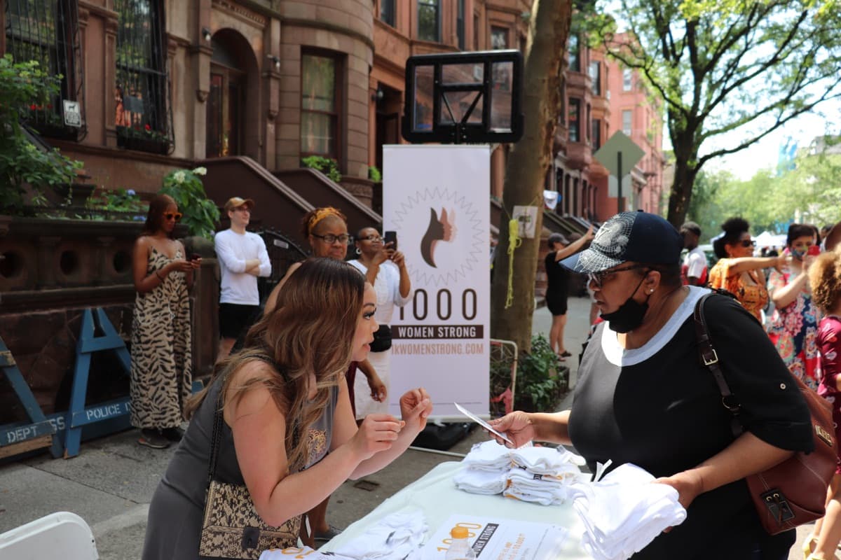 Two women talk across an outdoor table under a 1000 Women Strong banner at a street event.