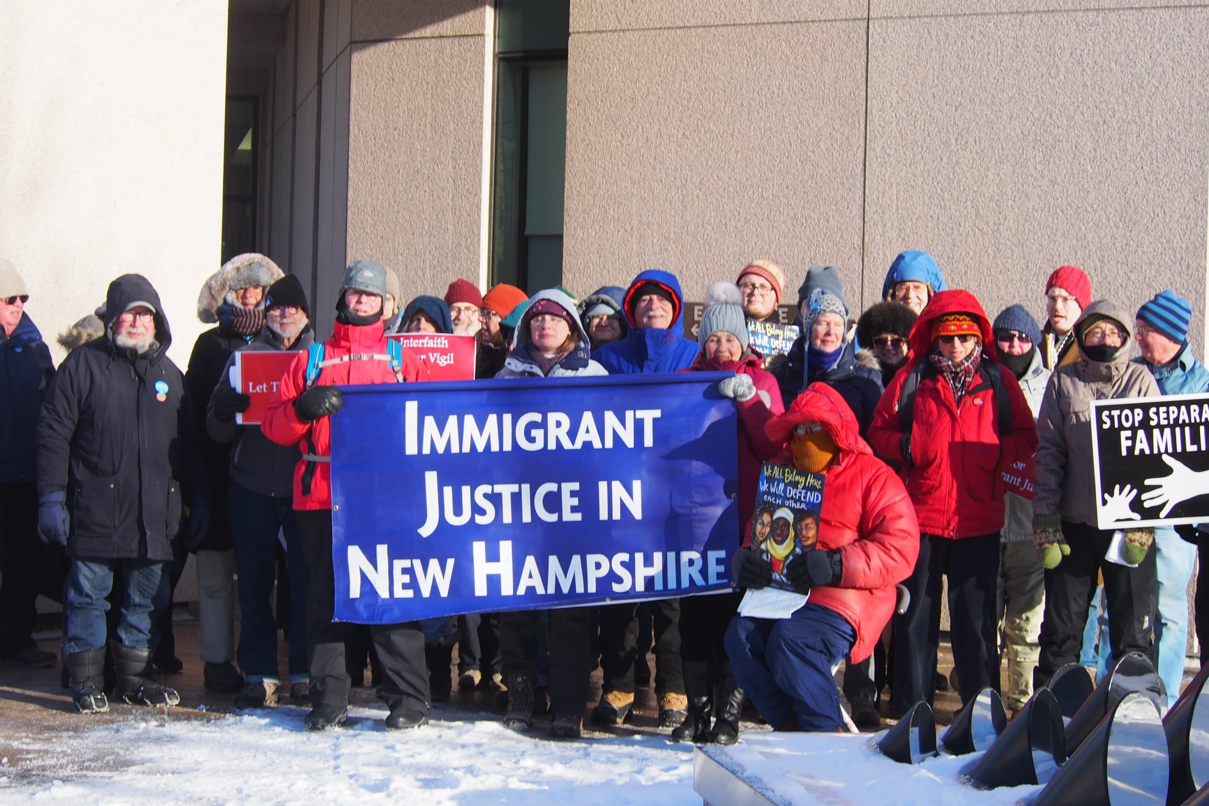 Granite State Interfaith supporters in winter coats hold an ‘Immigrant Justice in New Hampshire’ banner at a vigil in the snow