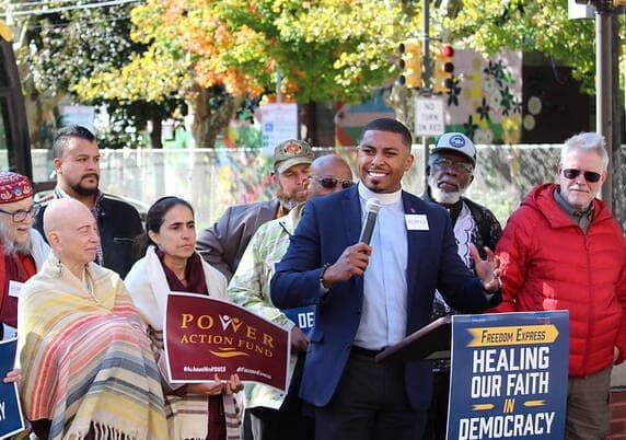 A POWER Action Fund speaker addresses a crowd of faith leaders and supporters holding signs outdoors.