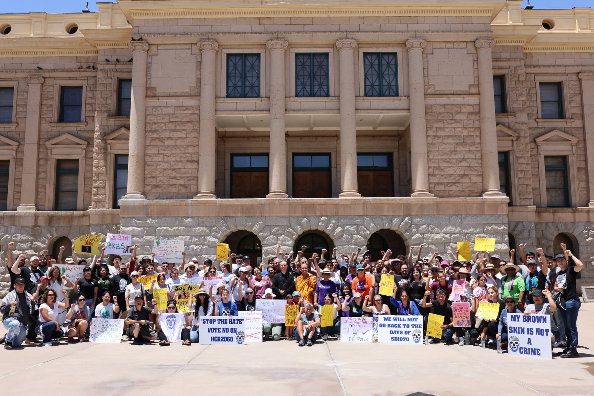 Dozens of people hold handmade signs, including “Stop the hate, vote no on HCR2060,” in front of the Arizona Capitol.