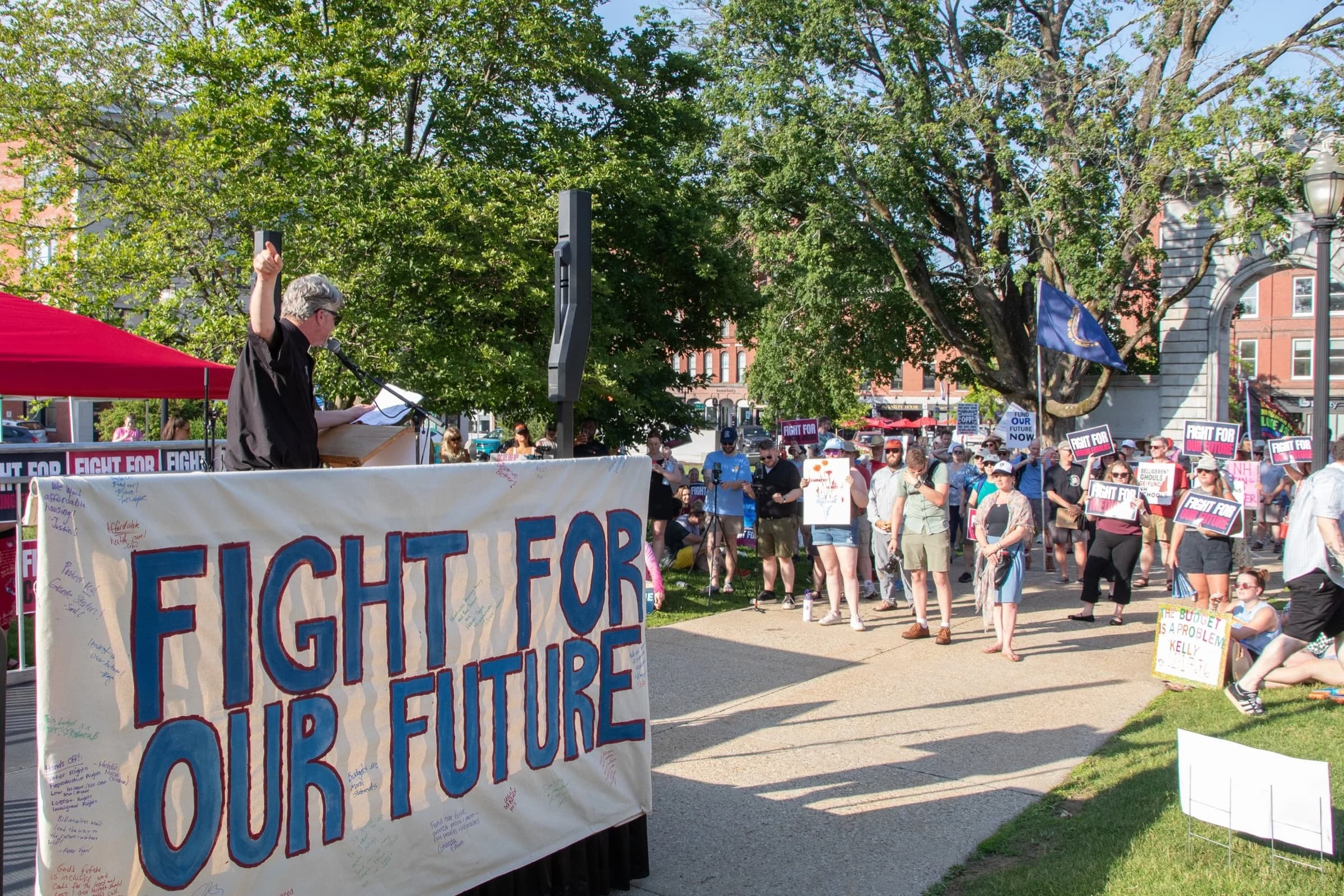 A speaker at a podium behind a hand-painted ‘Fight for Our Future’ banner addresses a crowd holding signs on the New Hampshire State House plaza in Concord