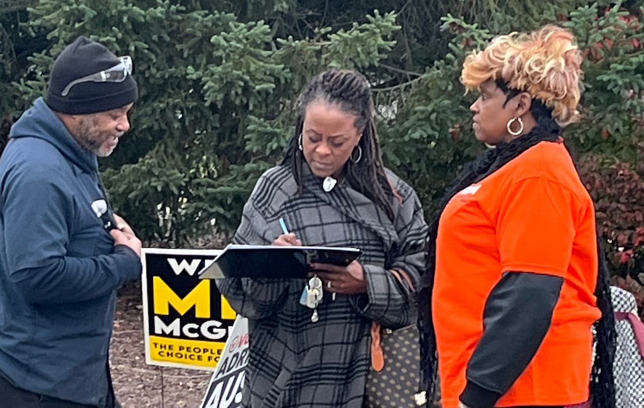 Three people talk outdoors as one writes on a clipboard, beside a People’s Choice yard sign.