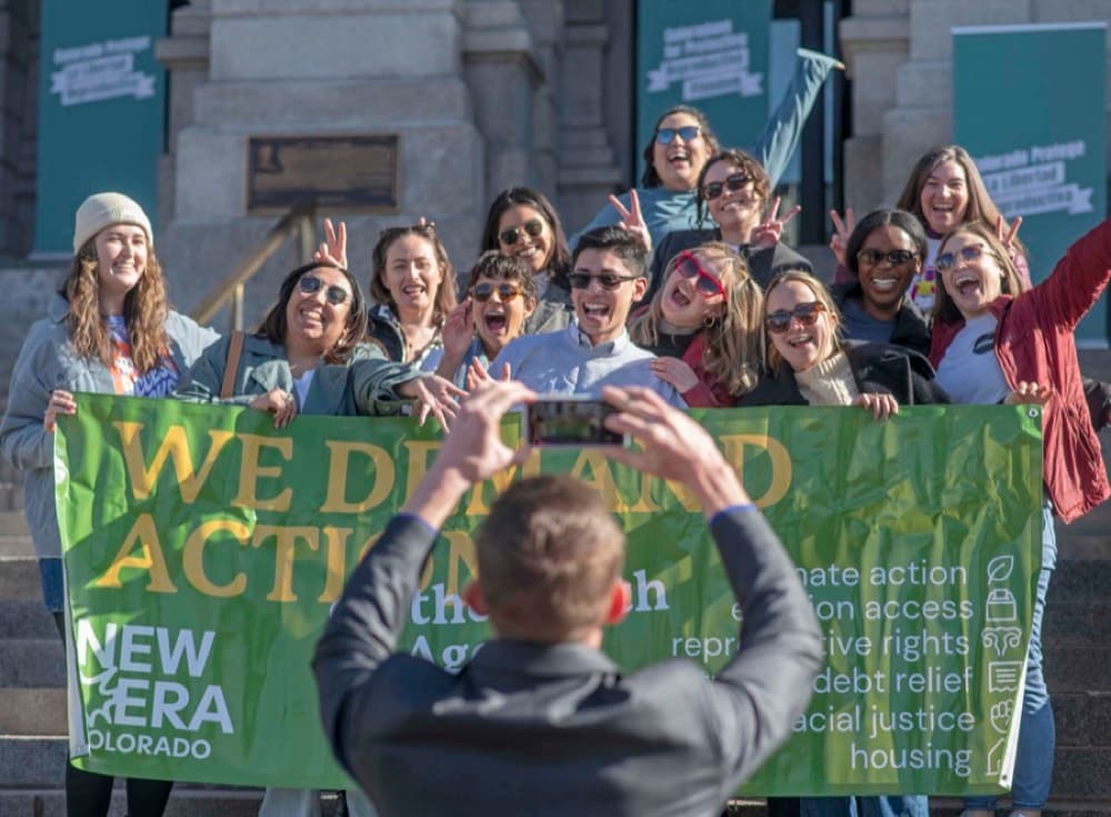 New Era Colorado members hold a “We demand action” banner behind a photographer.