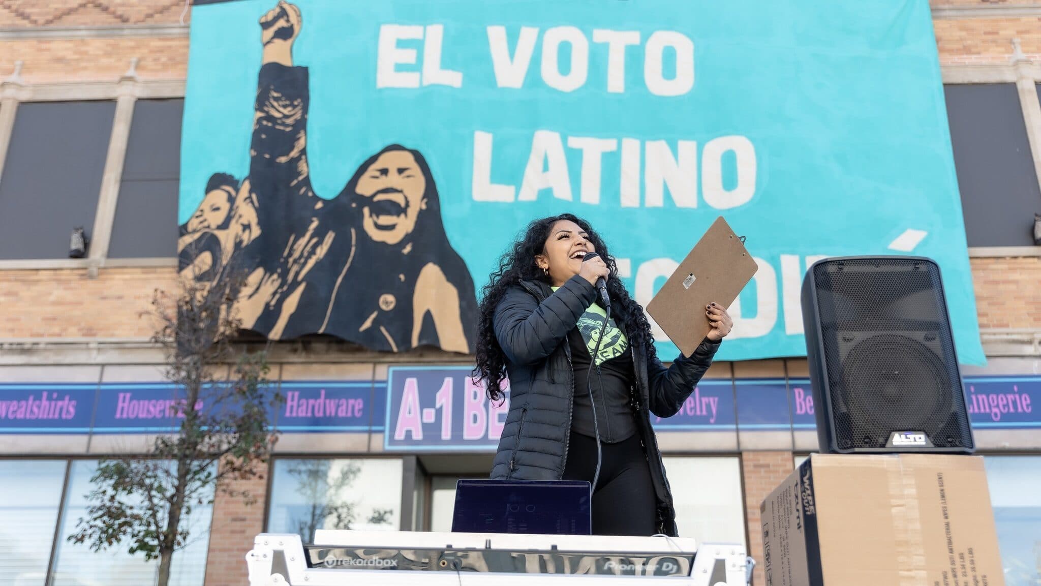 A Voces de la Frontera Action organizer speaks at a microphone in front of an El Voto Latino mural in Wisconsin.