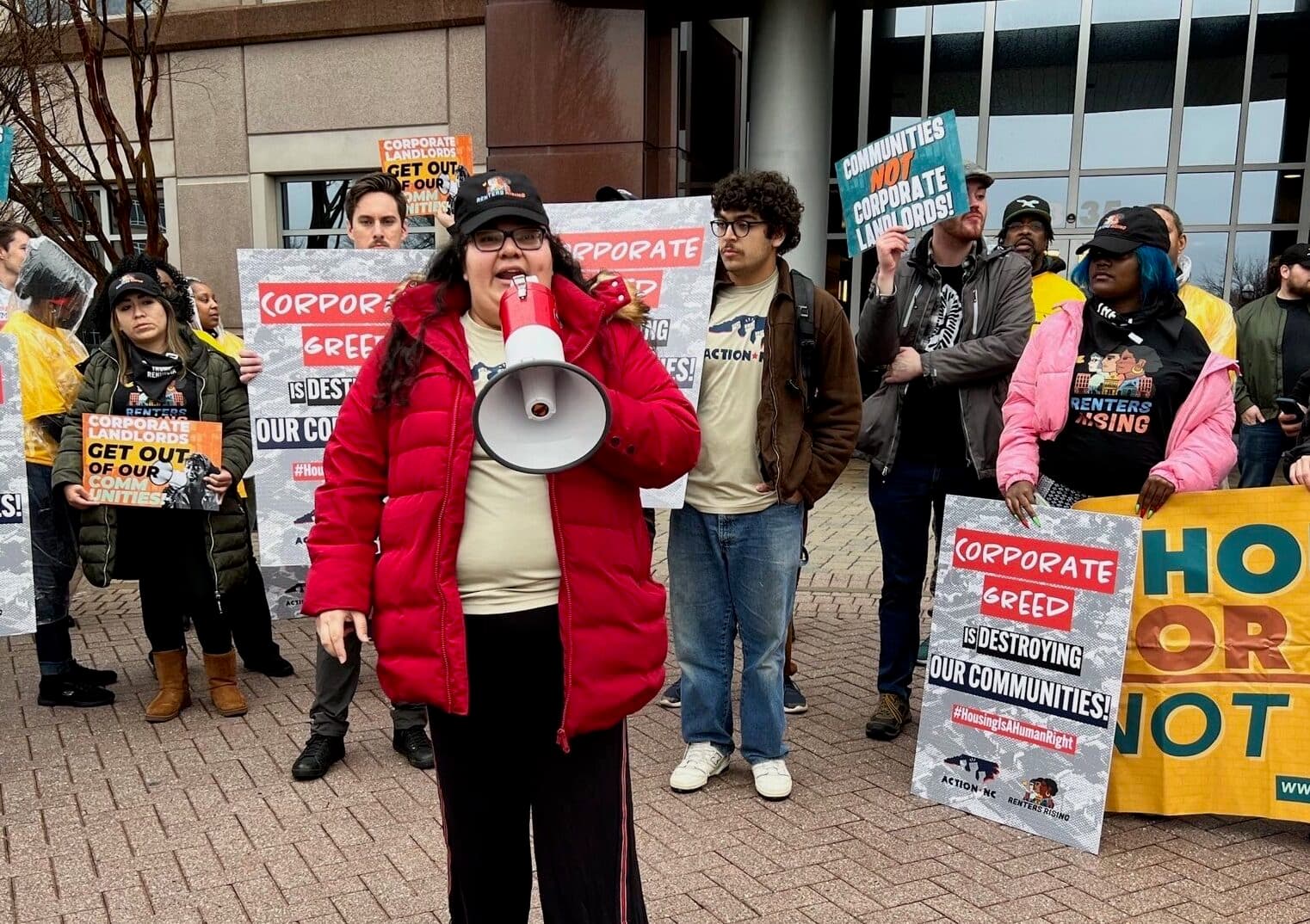 An Action NC organizer in a red coat speaks through a megaphone at a street protest, surrounded by members holding signs against corporate greed