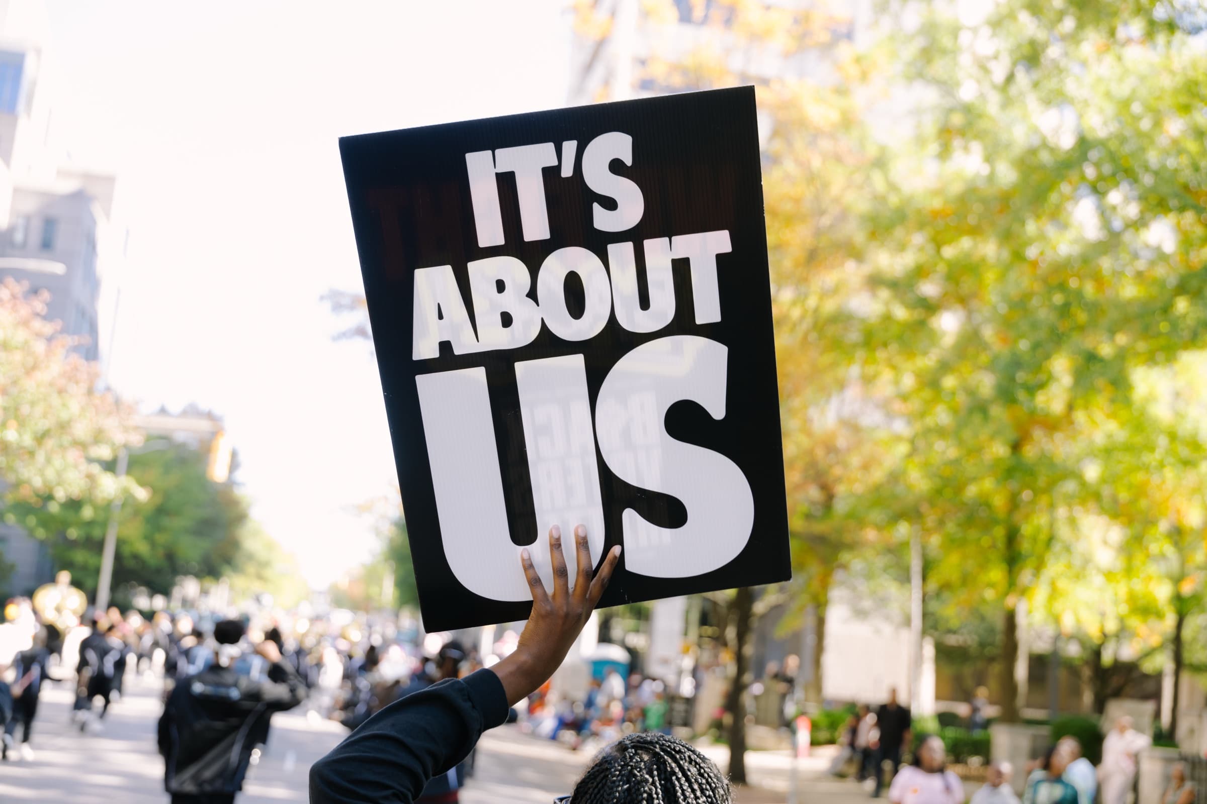 A marcher holds up a Black Voters Matter ‘It’s About Us’ sign above a crowd on a tree-lined city street