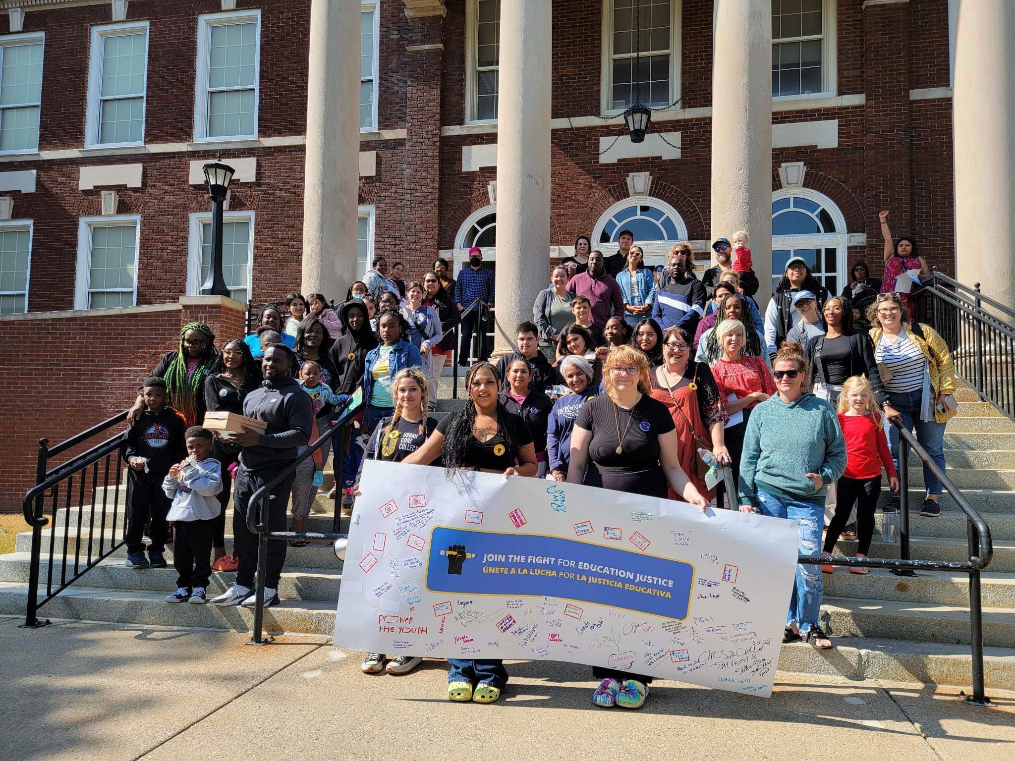 Dozens of Urban Core Collective members and families on the steps of a brick building with a signed ‘Join the Fight for Education Justice’ banner in Grand Rapids
