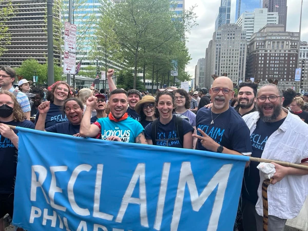 Reclaim Philadelphia members cheer behind a blue RECLAIM banner on a city street.
