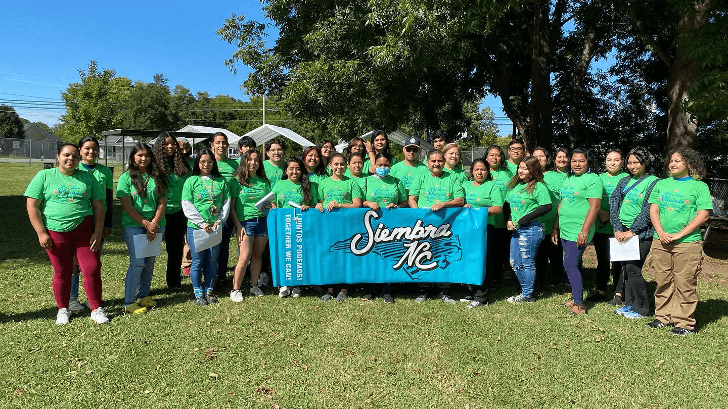 Siembra NC members in green shirts pose behind their banner on a lawn.