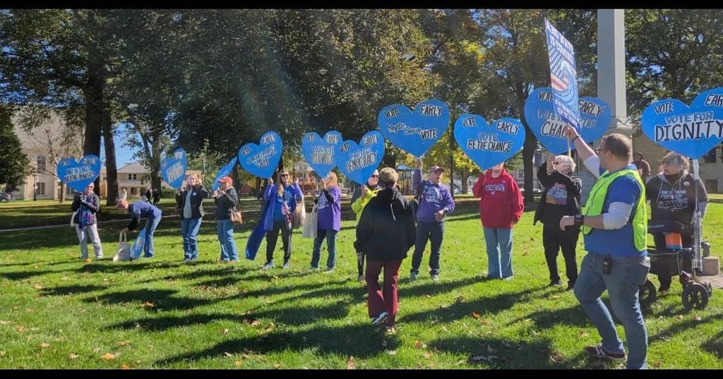 A group of people stands on a sunny lawn holding up blue heart-shaped signs that say Vote Early and Vote for Dignity.