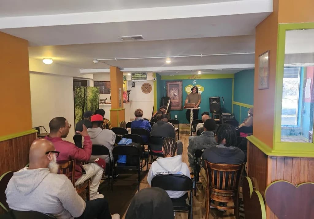 About two dozen people sit in rows of chairs in a brightly painted community room, facing a speaker standing at a podium with a laptop.